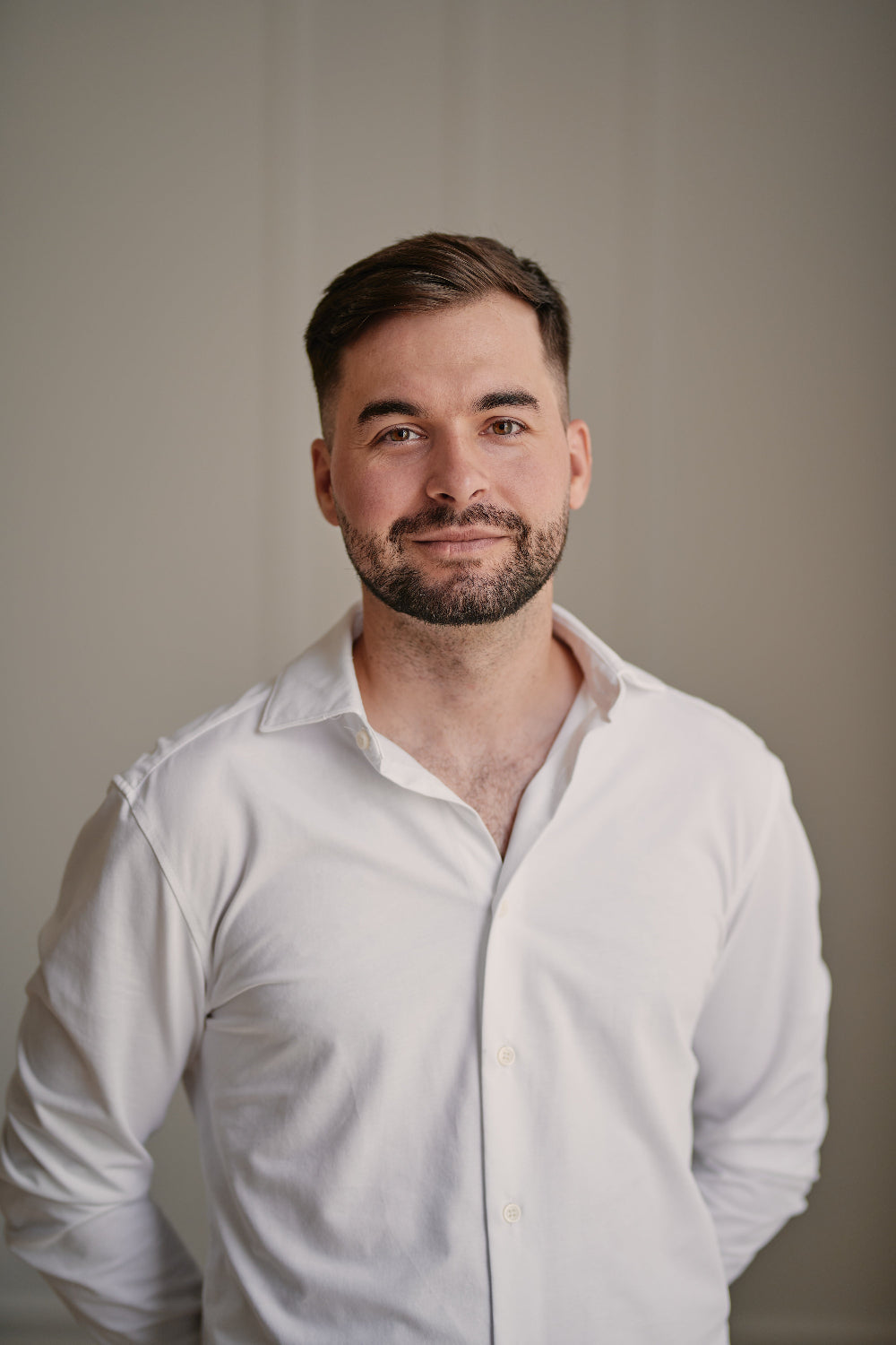 A man with short brown hair and a trimmed beard wearing a white long-sleeve button-down shirt, photographed against a plain warm gray background.